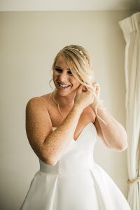 Smiling blonde bride putting on sparkly gold bridal teardrop earrings by J'Adorn Designs bridal jeweler for a classic Annapolis wedding at Chesapeake Bay Beach Club