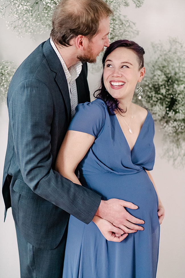 Maternity portrait featuring a smiling couple. The mother-to-be wears a slate grey dress and matching gemstone jewelry